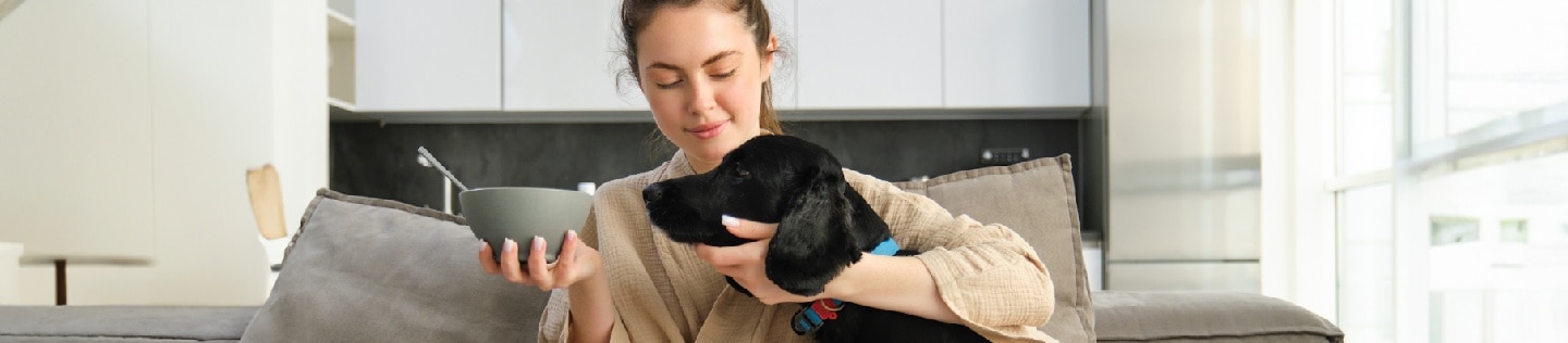 A black dog begging for food from a woman holding a bowl sitting on a sofa.