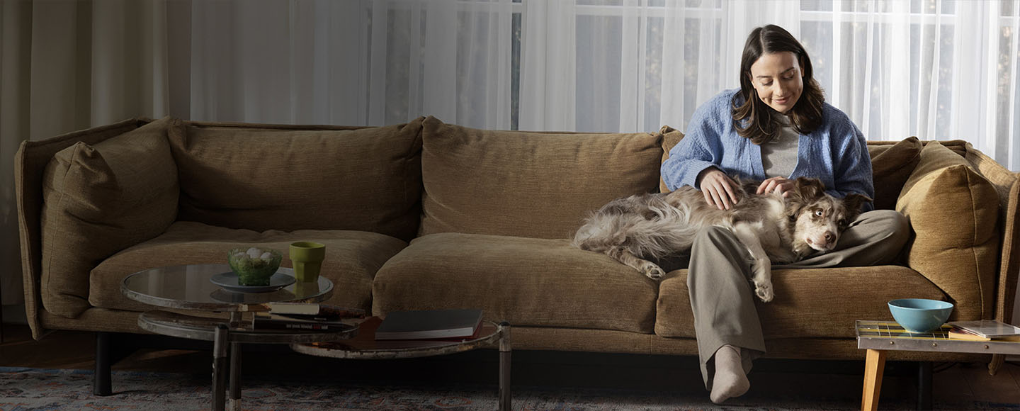 Woman sitting on a brown couch comfortably petting her dog resting on her lap.