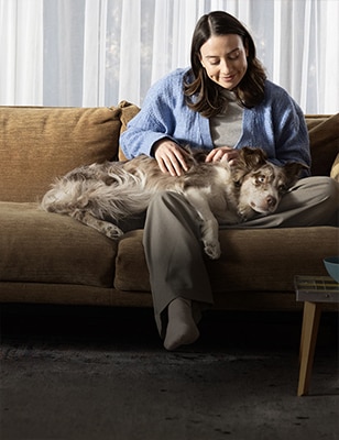 Woman sitting on a brown couch comfortably petting her dog resting on her lap.